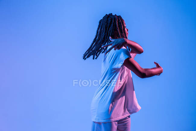 Dynamisches afroamerikanisches Teenager-Mädchen, das Bewegung macht, während es urbanen Tanz in Neonlicht vor blauem Hintergrund aufführt — Stockfoto