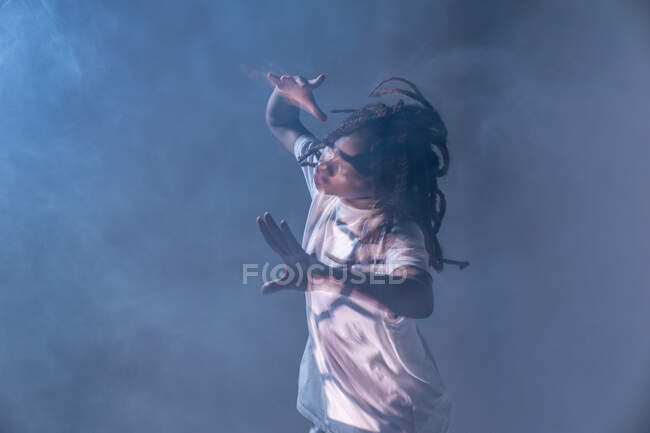 Dinámica afroamericana adolescente haciendo movimiento mientras realiza danza urbana en luz de neón contra fondo azul y humo - foto de stock
