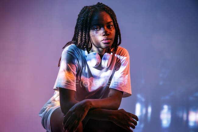 Full body of African American teenage girl with headphones sitting on neon lights in studio — Stock Photo