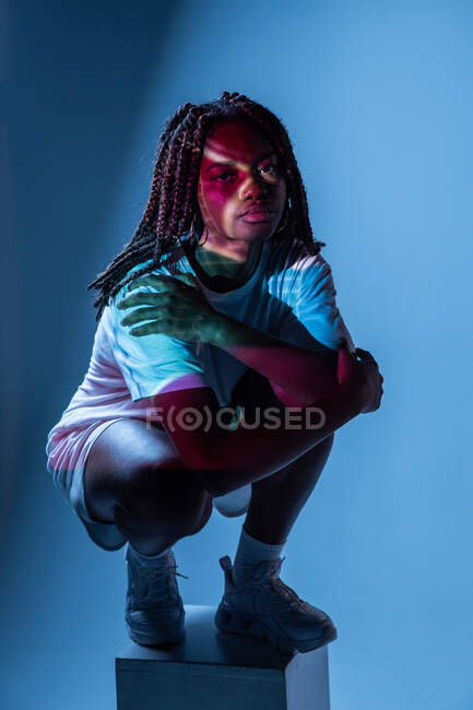 Full body of African American teenage girl sitting on pedestal in neon lights in studio — Stock Photo