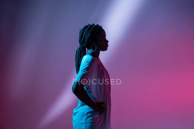 Vista lateral da adolescente afro-americana grave em fones de ouvido no pescoço de pé em estúdio com luzes brilhantes de néon — Fotografia de Stock