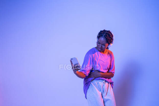 Serious African American teenage girl with smartphone standing in studio with bright glowing neon lights — Stock Photo