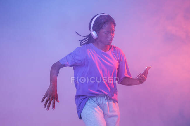 Chica adolescente afroamericana seria con teléfono inteligente en auriculares de pie en el estudio con luces de neón brillantes y humo - foto de stock