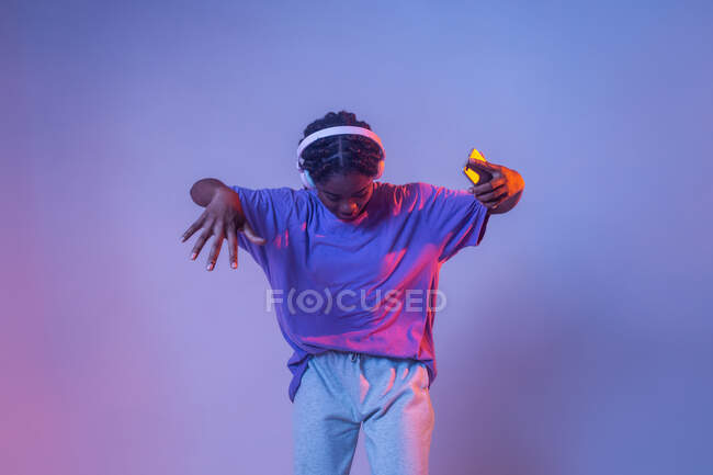 Serious African American teenage girl with smartphone in headphones standing in studio with bright glowing neon lights — Stock Photo