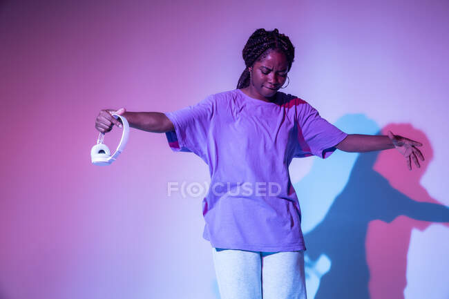 Cuerpo completo de chica adolescente afroamericana con auriculares en la mano realizando movimiento de danza urbana en estudio brillante - foto de stock