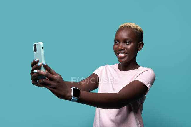 Expresiva hermosa mujer afroamericana con manicura brillante tomando autorretrato en el teléfono inteligente contra fondo azul - foto de stock