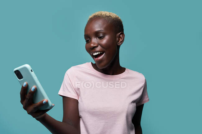 Expressive belle femme afro-américaine aux cheveux courts et à la manucure lumineuse naviguant sur smartphone sur fond bleu — Photo de stock
