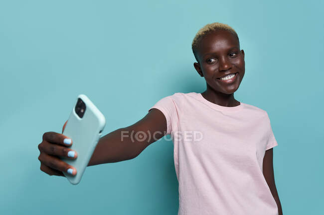 Expressive belle femme afro-américaine avec manucure lumineuse prenant autoportrait sur smartphone sur fond bleu — Photo de stock
