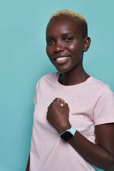 Happy African American female with trendy hairstyle demonstrating smartwatch while standing against blue background and looking at camera - foto de stock