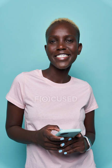 Expressive beautiful African American female with short hair and bright manicure browsing on smartphone while looking at camera against blue background — Stock Photo