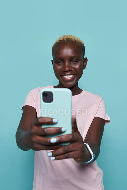 Expresiva hermosa mujer afroamericana con manicura brillante tomando autorretrato en el teléfono inteligente contra fondo azul - foto de stock