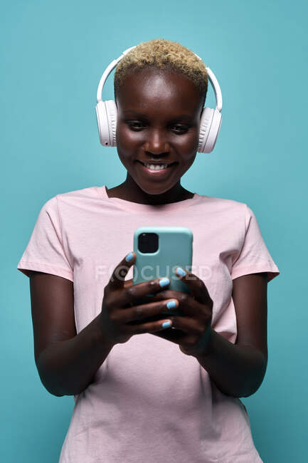 Fröhliche afrikanisch-amerikanische Frau zahm lächelnd, während sie Musik im Kopfhörer vor blauem Hintergrund hört — Stockfoto
