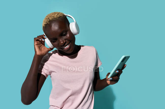 Cheerful African American female toothy smiling while dancing and listening to music in headphones against blue background — Fotografia de Stock