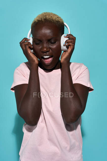 Cheerful African American female smiling and singing while listening to music in headphones against blue background — Foto stock