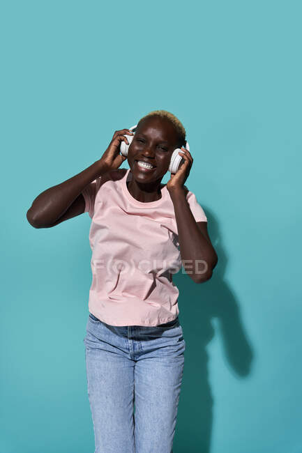 Cheerful African American female toothy smiling looking at camera listening to music in headphones against blue background - foto de stock