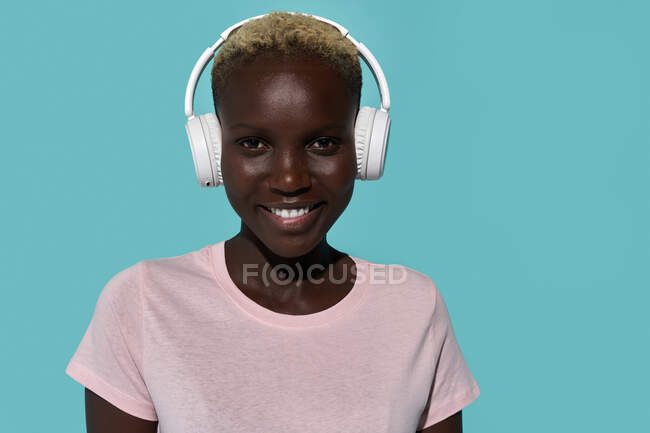 Serene beautiful African American female smiling looking at camera while listening to music in headphones against blue background - foto de stock