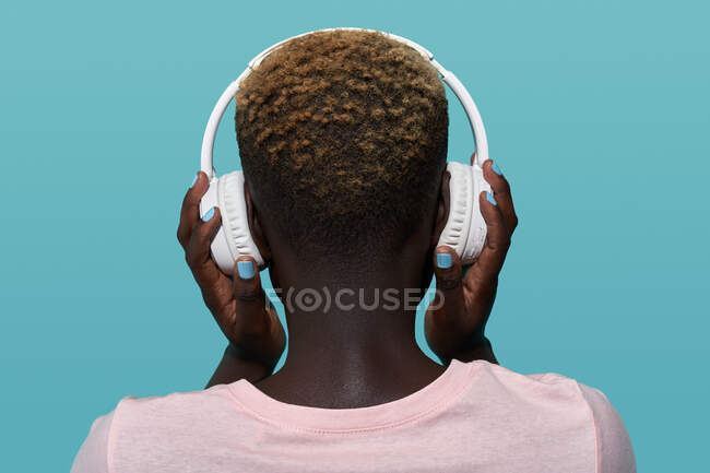 Back view of faceless African American female with short hair listening to music in headphones while standing against blue background — Foto stock