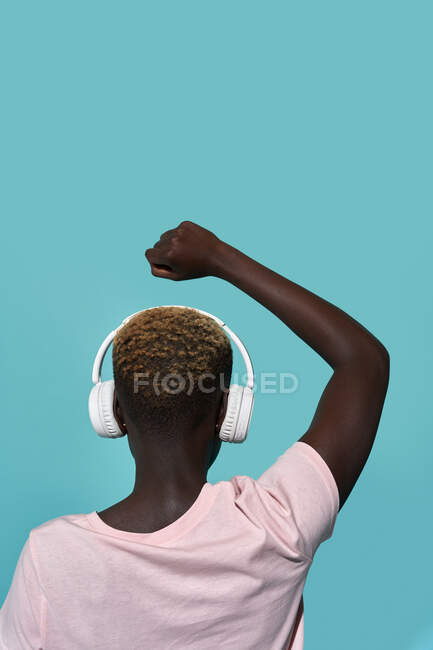 Back view of faceless African American female with raised arm and fist closed listening to music in headphones while standing against blue background — Fotografia de Stock