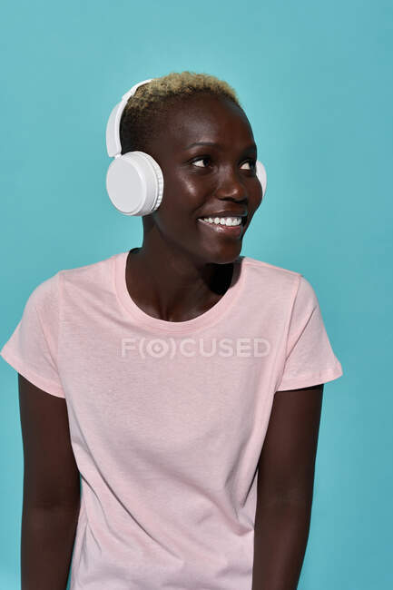 Cheerful African American female toothy smiling looking away listening to music in headphones against blue background — Foto stock