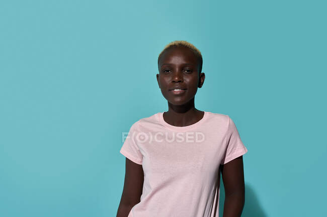 Спокійна африканська жінка, яка посміхається, дивлячись на камеру, що стоїть навпроти синього фону у студії. — стокове фото