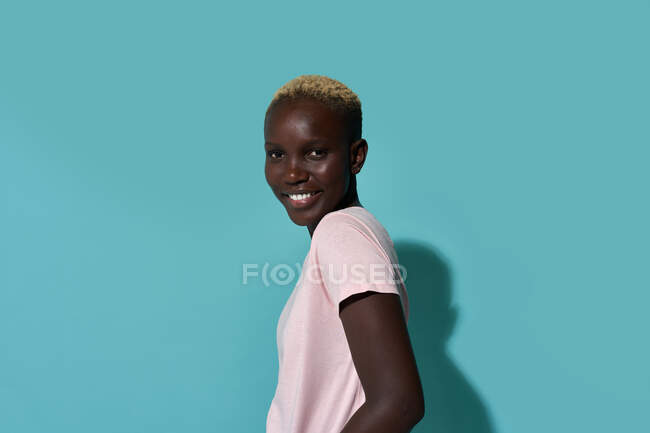 Side view of serene cute beautiful African American female smiling looking at camera standing against blue background in a studio — Stock Photo