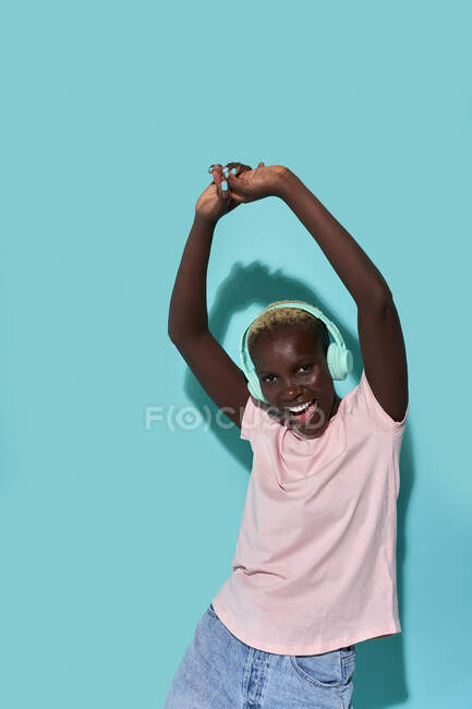 Cheerful African American female toothy smiling with arms raised dancing looking at camera while listening to music in headphones against blue background — Stock Photo