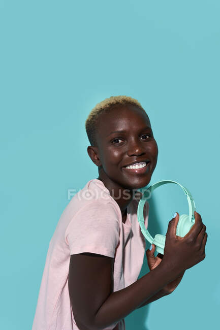 Side view of cheerful African American female toothy smiling looking at camera listening to music in headphones against blue background - foto de stock