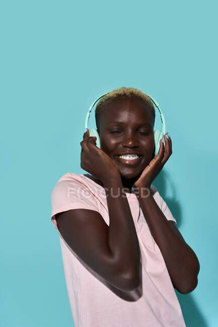 Cheerful African American female toothy smiling with eyes closed listening to music in headphones against blue background - foto de stock
