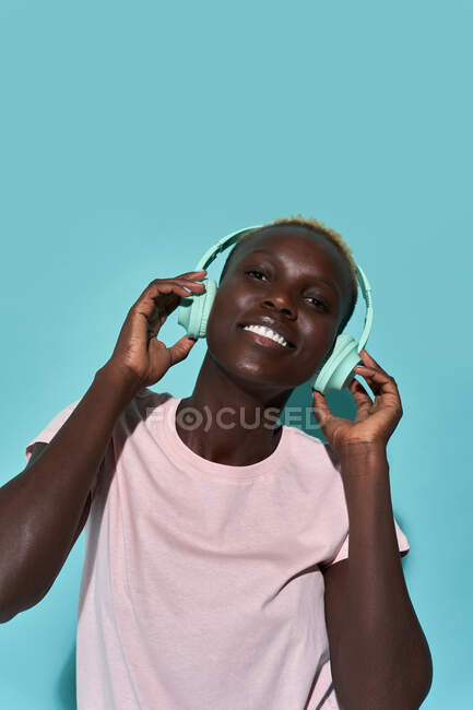 Cheerful African American female toothy smiling looking at camera listening to music in headphones against blue background — Foto stock
