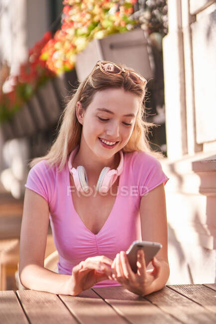 Fröhliche Frau mit drahtlosen Kopfhörern im Nacken, die mit dem Handy im Internet surft, sitzt am Tisch in einem Straßencafé in Madrid — Stockfoto