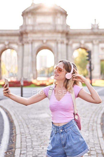 Весела жінка в повсякденному одязі і сонцезахисних окулярах робить автопортрет, слухаючи музику на божевільній вулиці. — стокове фото