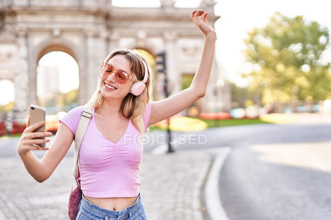 Весела жінка в повсякденному одязі і сонцезахисних окулярах робить автопортрет, слухаючи музику на божевільній вулиці. — стокове фото
