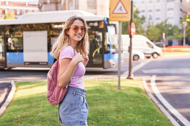 Vista lateral de fêmea positiva com mochila à espera de transporte na calçada durante viagem em Madrid — Fotografia de Stock