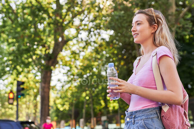 Vue latérale d'une jeune femme joyeuse avec sac à dos et bouteille d'eau debout dans un parc verdoyant par une journée ensoleillée à Madrid — Photo de stock