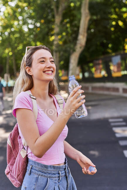 Seitenansicht einer fröhlichen jungen Frau mit Rucksack und Wasserflasche, die im grünen Park an einem sonnigen Tag in Madrid steht — Stockfoto