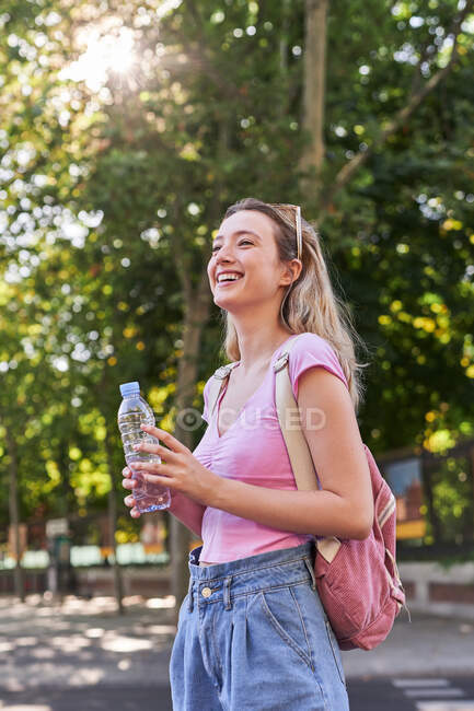 Боковой вид веселой молодой женщины с рюкзаком и бутылкой воды, стоящей в зеленом парке в солнечный день в Мадриде — стоковое фото