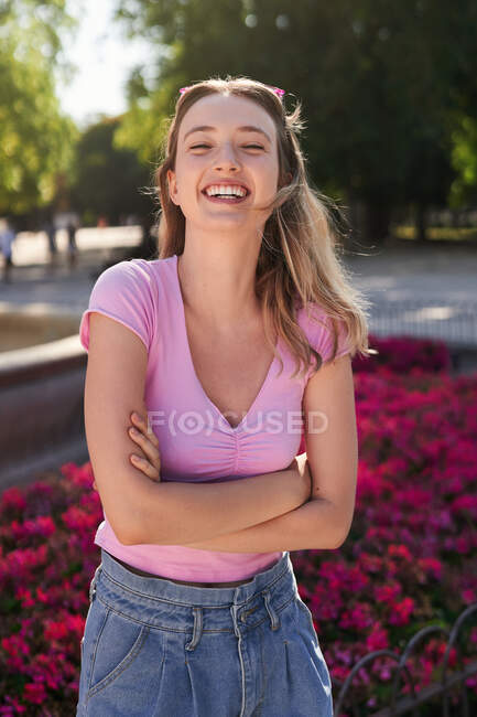 Femme souriante avec bras croisés regardant caméra dans le parc avec des arbres verts et des fleurs rouges à Madrid — Photo de stock