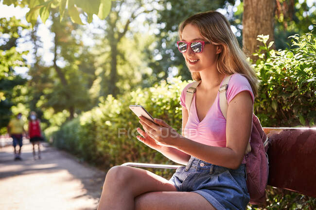 Seitenansicht einer fröhlichen Frau mit Handy, die während einer Reise in Madrid im grünen Park sitzt — Stockfoto