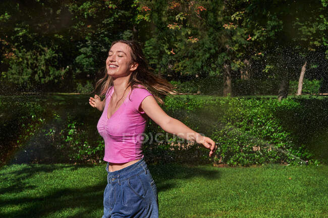 Весела жінка з піднятими руками, стоячи в бризках в сонячному парку — стокове фото