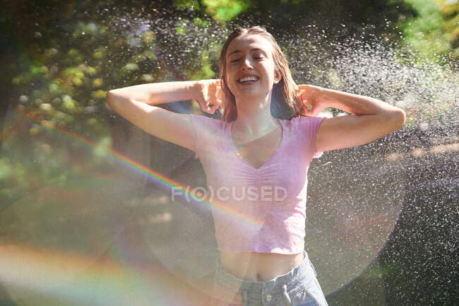 Femme joyeuse avec les mains sur la tête debout dans des éclaboussures dans un parc ensoleillé — Photo de stock
