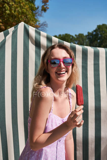 Mujer alegre en vestido de verano de pie con paleta y disfrutando de un día soleado en Madrid - foto de stock