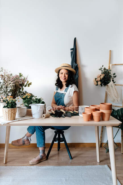 Young female horticulturist in straw hat sitting near flowers on table with assorted tools at home — Fotografia de Stock