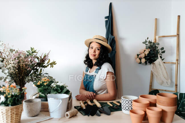 Young female horticulturist in straw hat sitting near flowers on table with assorted tools at home - foto de stock