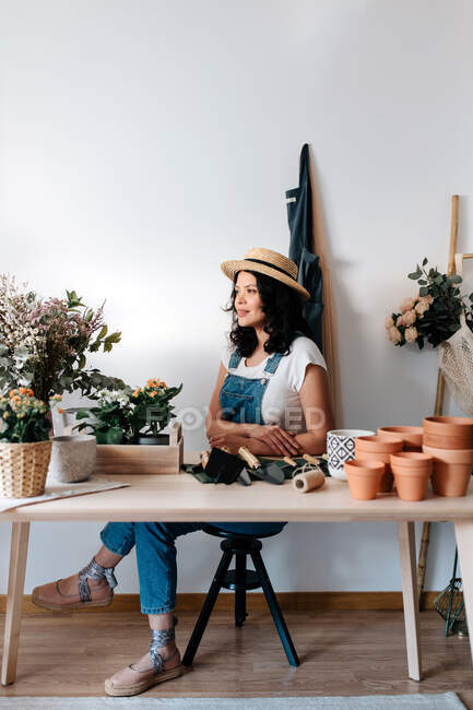 Young female horticulturist in straw hat sitting near flowers on table with assorted tools at home — Foto stock