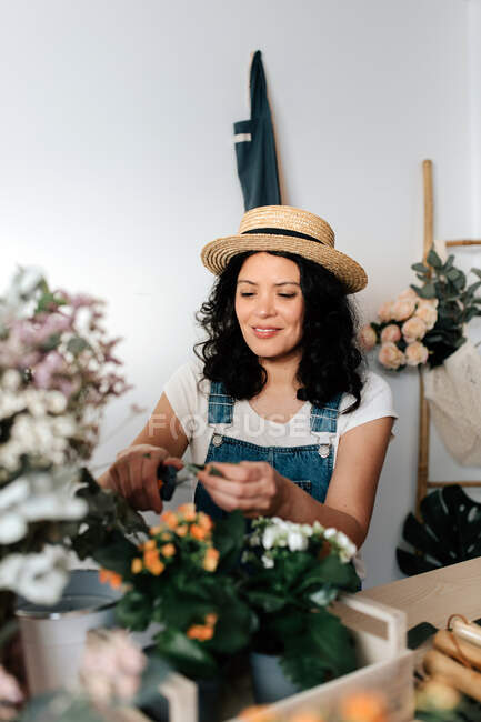 Молода жінка садівниця в солом'яному капелюсі створює букет на столі з різноманітними інструментами вдома — стокове фото