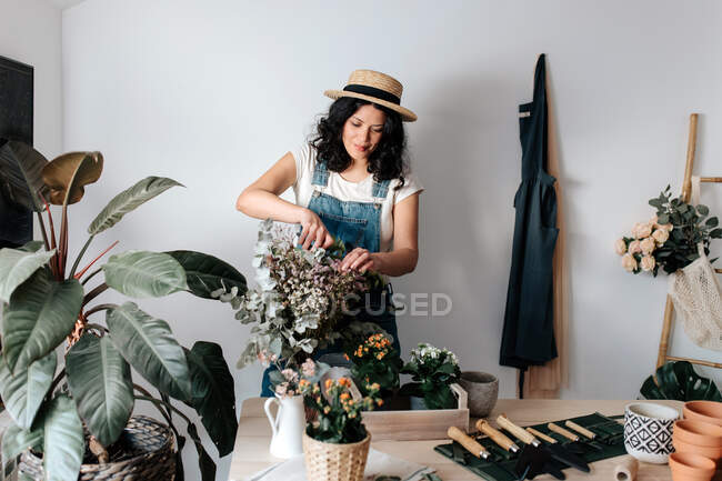 Junge Gärtnerin mit Strohhut schneidet Pflanzenblätter in der Nähe von Topfblumen auf dem Tisch mit verschiedenen Werkzeugen zu Hause — Stockfoto