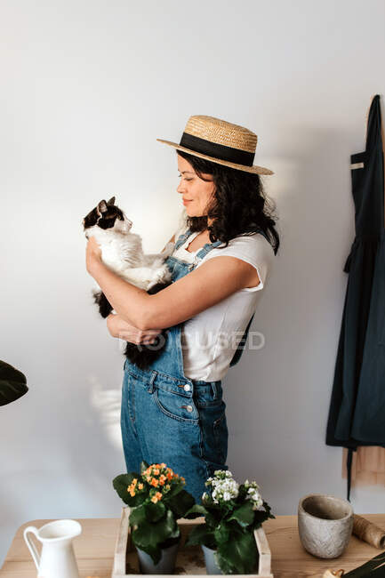 Side view of young glad female horticulturist stroking and embracing fluffy cat at table with blooming potted flowers in house — Fotografia de Stock