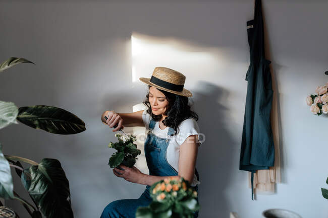 Jeune jardinière en tenue décontractée à l'aide d'un instrument de jardinage sur fleurs fleuries de plantes en pot à la maison — Photo de stock