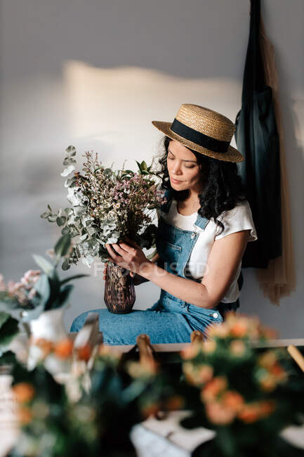 Young female horticulturist in straw hat creating bouquet on table with assorted tools at home — Foto stock