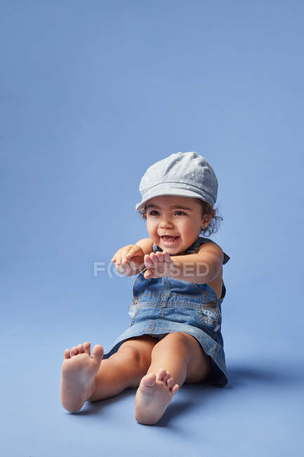 Charmant fröhliches Barfußkind in Jeanskleid und Hut mit lockigem Haar, das beim Spielen auf blauem Hintergrund wegschaut — Stockfoto